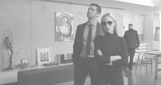 [Image Description: A man and a woman stand together in a dining room. The man stand with his suit jacket open looking up. The woman stands to his right with her arms crossed and her sunglasses on. She has her hip cocked out to the right and she does not look happy.]